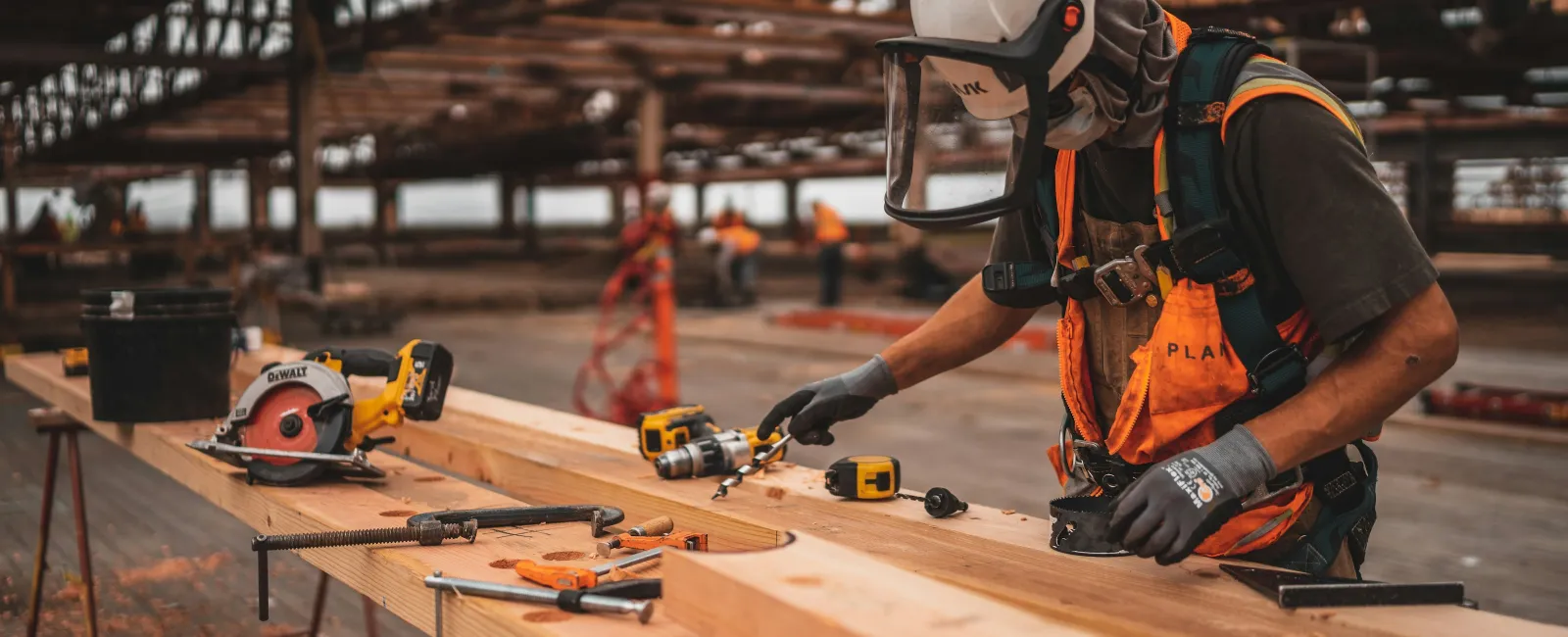 Construction worker wearing safety gear measuring wooden planks in a large open industrial building.