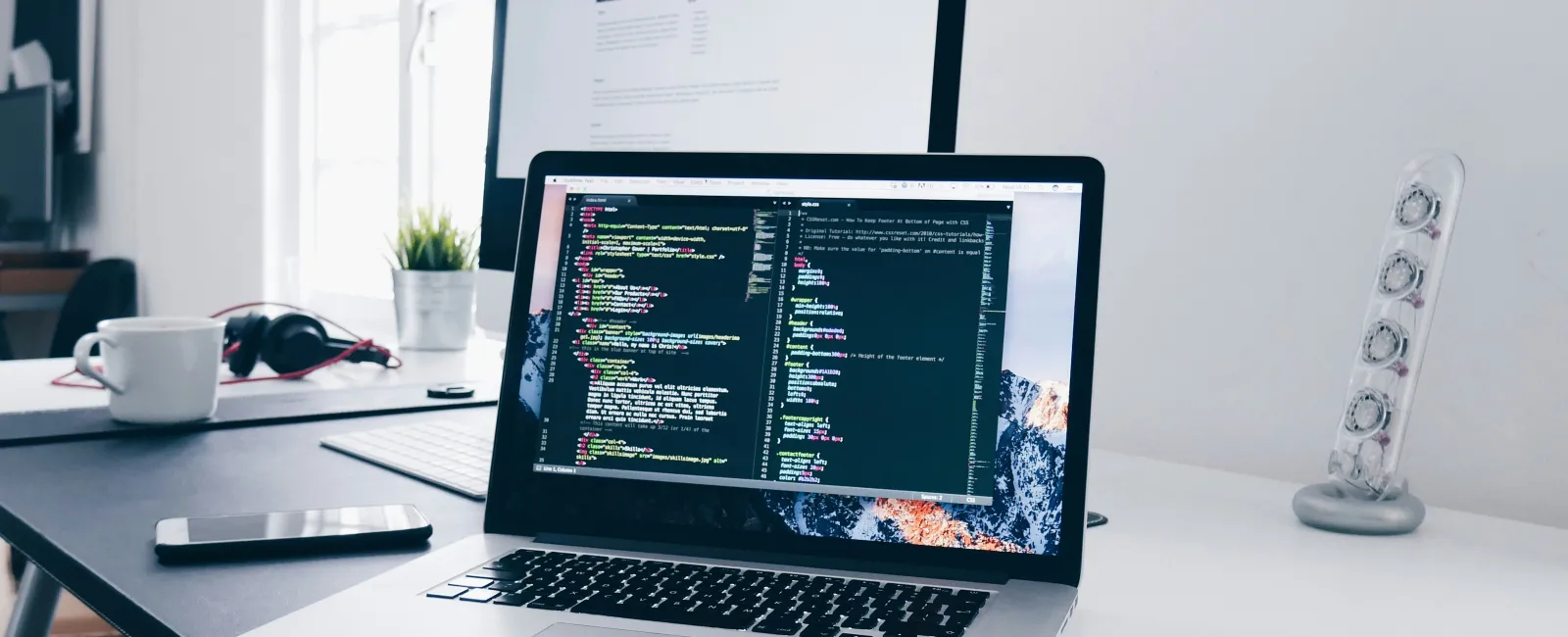Laptop on desk displaying colorful coding text with a large monitor, smartphone, coffee cup, and desk lamp nearby.