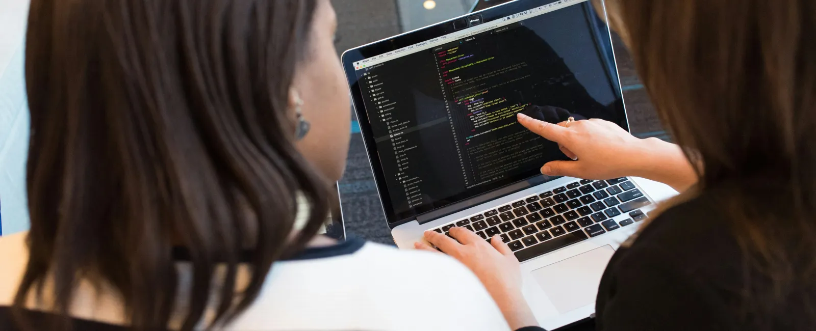 Two women collaborating on coding or programming on a laptop screen in a modern workspace.