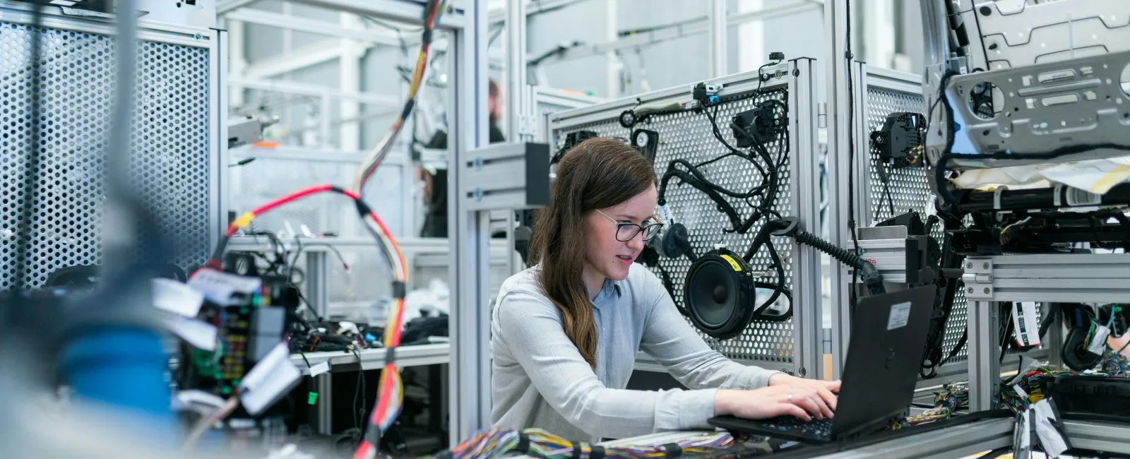 Female engineer working on laptop inside a modern automotive testing lab with wiring and car parts