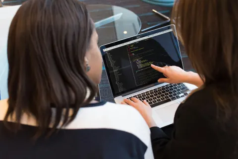 Two women collaborating on coding or programming on a laptop screen in a modern workspace.