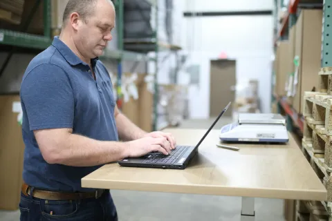 Man using laptop on a standing desk in a warehouse surrounded by pallets and boxes.