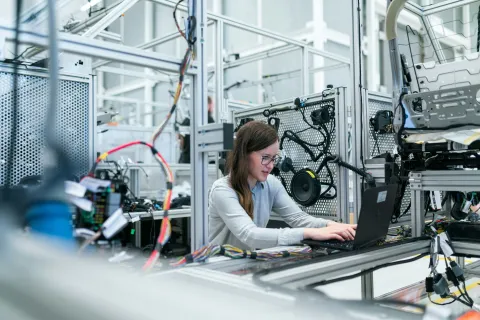 Female engineer working on laptop inside a modern automotive testing lab with wiring and car parts