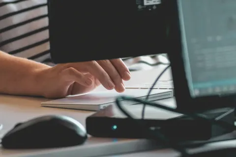 Person typing on a white keyboard at a desk with computer monitors and a mouse nearby